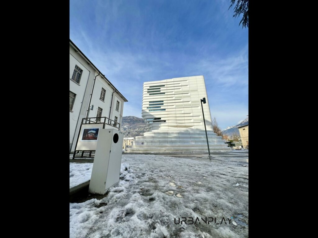 Cestino lungo il percorso pedonale con edificio universitario e montagne sullo sfondo