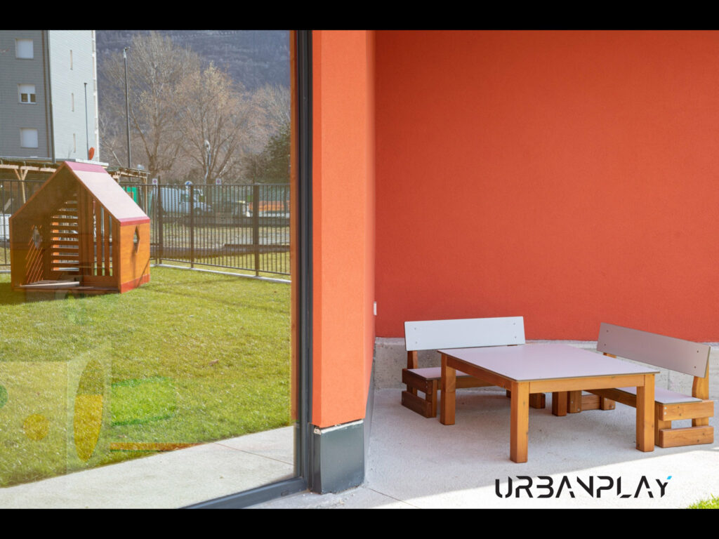 Area esterna coperta con tavolo, panche e casetta in legno nel giardino del nido