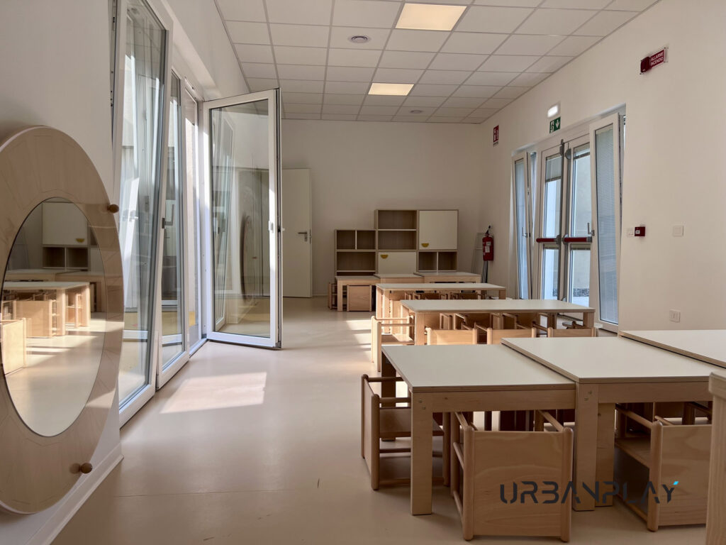 Aula luminosa con tavoli, sedie, specchio e arredi bassi per bambini