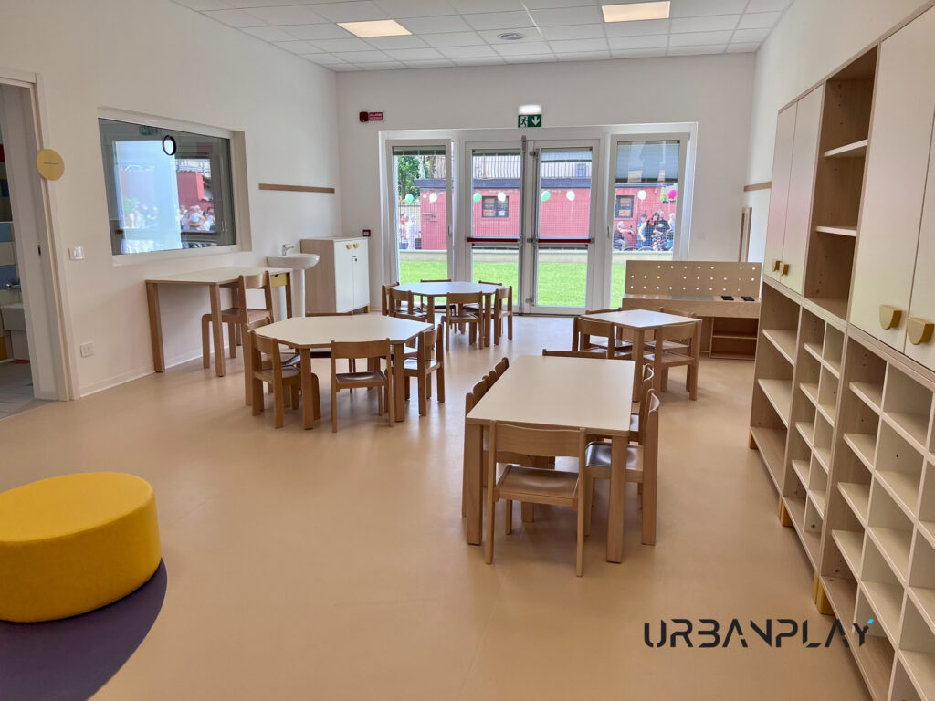 Aula arredata con tavoli, sedie, armadiature, scaffali aperti e mobile gioco